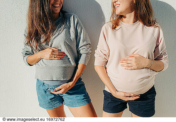 Two young pregnant woman holding belly while smiling at each other