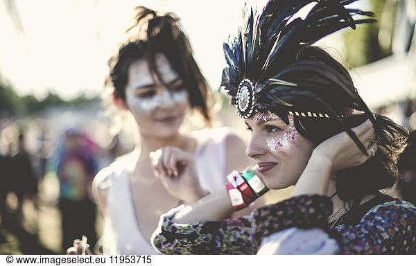 Two young women at a summer music festival faces painted  wearing feather headdress.