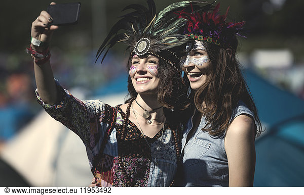 Two young women at a summer music festival faces painted  wearing feather headdress  taking selfie with smartphone.