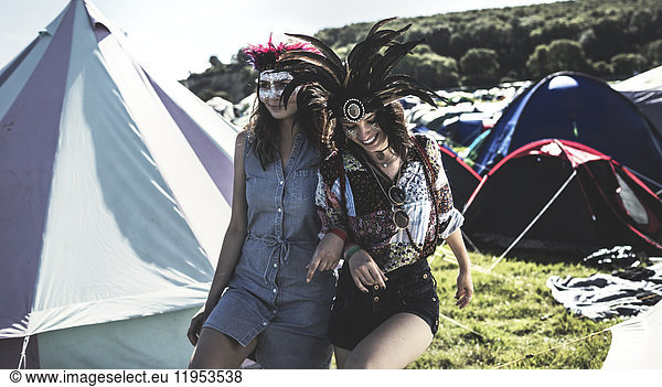 Two young women at a summer music festival faces painted  wearing feather headdresses  walking arm in arm in between tents.
