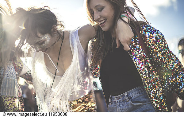 Two young women at a summer music festival wearing sequins with painted faces laughing and dancing.