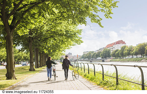 Two young women walking along river and pushing bikes