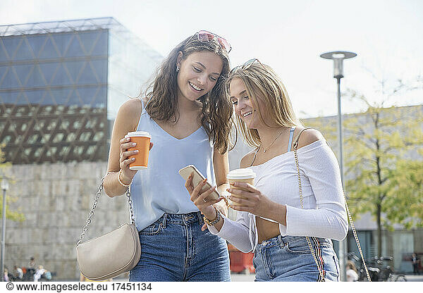 Two young women with coffee cups watching on mobilphone in City