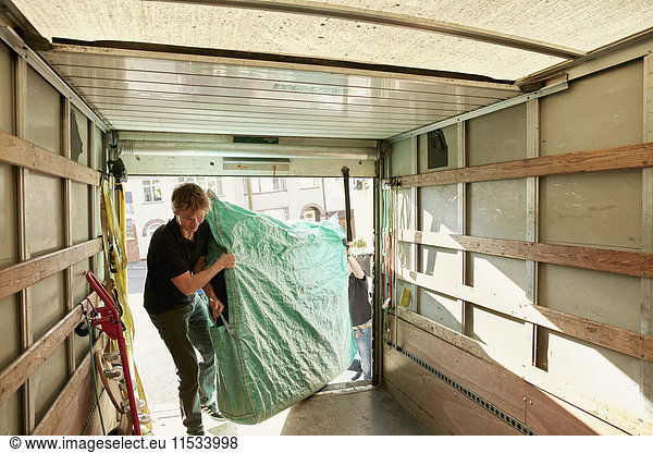Umzugsunternehmen. Ein Mann hebt ein mit grünem Plastik überzogenes Möbelstück in einen Umzugswagen.