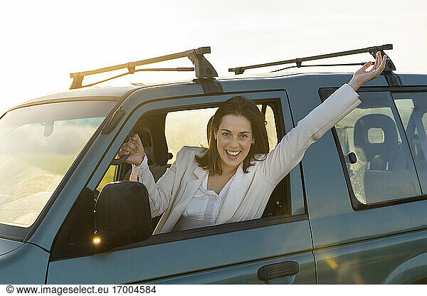 Unbekümmerte junge Frau lehnt sich bei Sonnenuntergang mit erhobenen Armen aus dem Autofenster