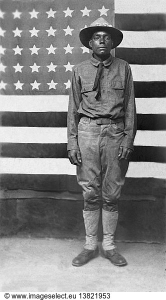 United States: c. 1916 An African American soldier in uniform in front of an American flag.