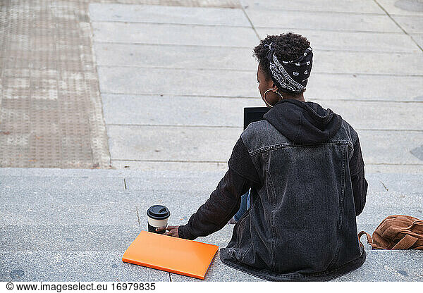 University female african student drinking coffee studying on her laptop sitting on stairs backwards outside on campus. College life concept.