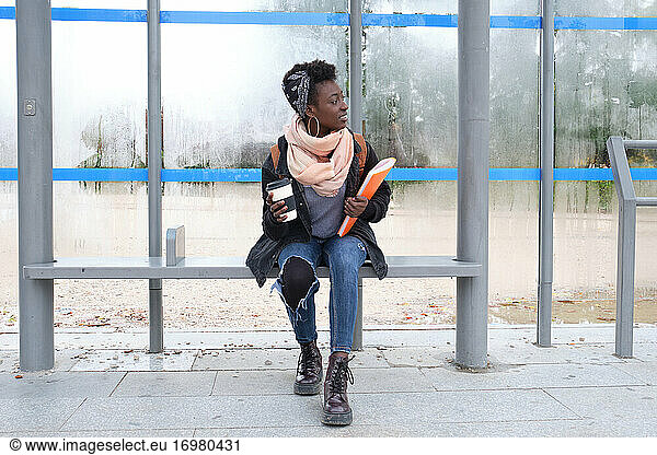 University female african student waiting for a bus holding a coffee cup and a folder on campus. College life concept.