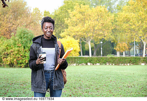 University female african student with backpack  folder and a cup of coffee outside on campus. College life concept.