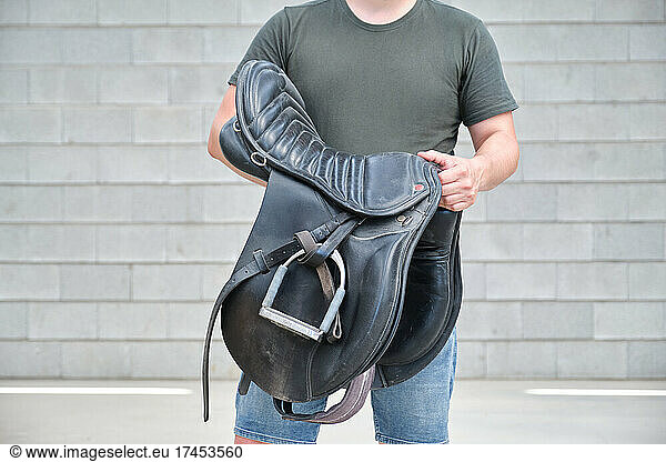 Unrecognizable man holding a saddle over gray brick background.