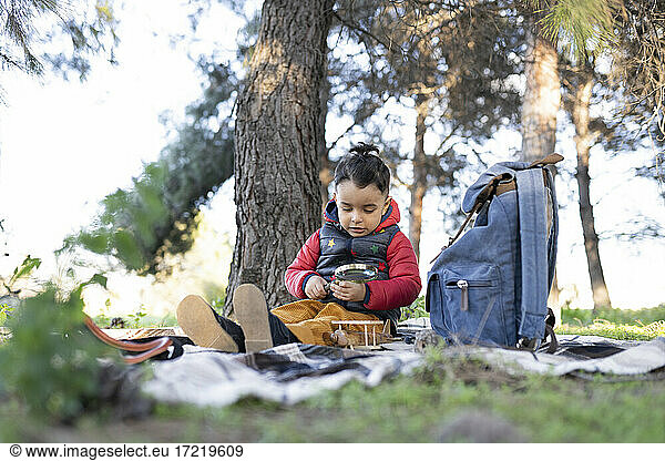 Unschuldiger Junge spielt mit einem Vergrößerungsglas an einem Rucksack in einem öffentlichen Park