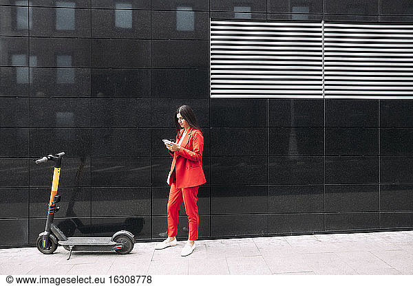 Unternehmerin in roter Geschäftskleidung  die ihr Smartphone benutzt  während sie vor einem schwarzen Gebäude in der Stadt steht