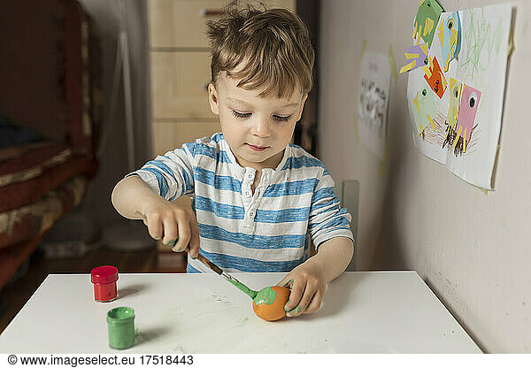 Upper body of blonde boy painting orange easter egg with green p