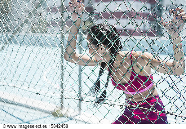 Upper body of female athlete leaning on court fence while resting