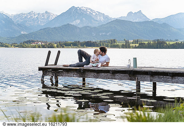 Vater  der seine Tochter küsst  während er auf einem Steg über dem See vor den Bergen sitzt