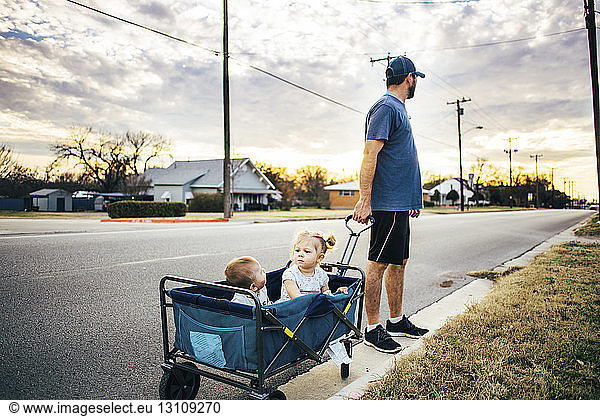 Vater hält Wagen mit Kinderwagen  in dem Kinder auf der Straße sitzen