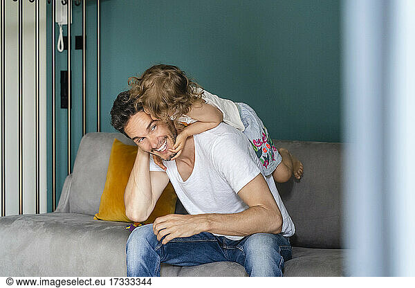 Vater nimmt verspielte Tochter huckepack  während er auf dem Sofa im Wohnzimmer sitzt