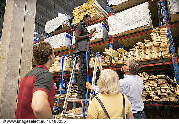 Verkäuferin steht auf einer Leiter und gestikuliert  während sie mit Kunden in einem Baumarkt spricht