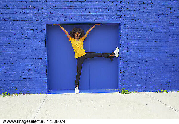 Verspielte Frau mit erhobenen Händen an blauer Wand lehnend