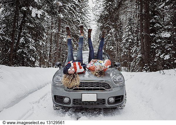 Verspielte Freundinnen in voller Länge  die im Winter mit erhobenen Füßen auf der Motorhaube eines Autos liegen