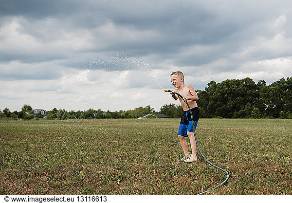 Verspielter Junge in voller Länge ohne Hemd  der mit einem Gartenschlauch auf einem Grasfeld vor bewölktem Himmel im Hinterhof spielt