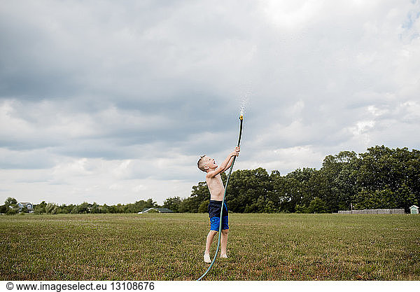 Verspielter Junge in voller Länge ohne Hemd  der mit einem Gartenschlauch Wasser auf einem Grasfeld gegen den bewölkten Himmel im Hinterhof spritzt