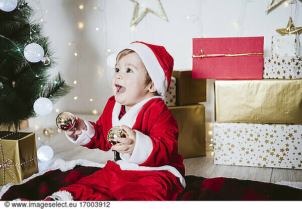 Verspielter kleiner Junge im Weihnachtsmannkostüm  der mit einer Weihnachtskugel spielt  während er zu Hause auf einer Decke sitzt