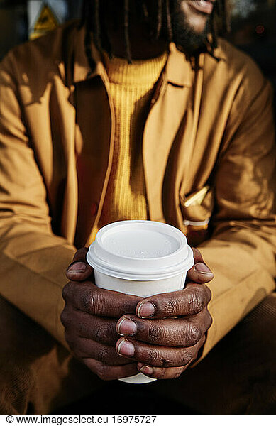 vertical shot of an african guy holding a paper cup of coffee with both hands. focus on the cup.