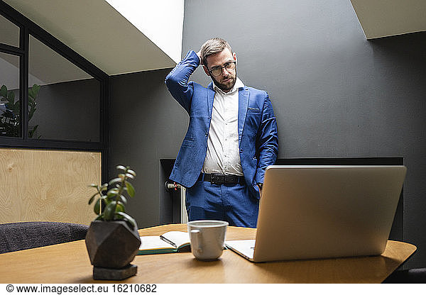 Verwirrter Geschäftsmann  der sich am Kopf kratzt  während er den Laptop auf dem Schreibtisch im Kreativbüro betrachtet