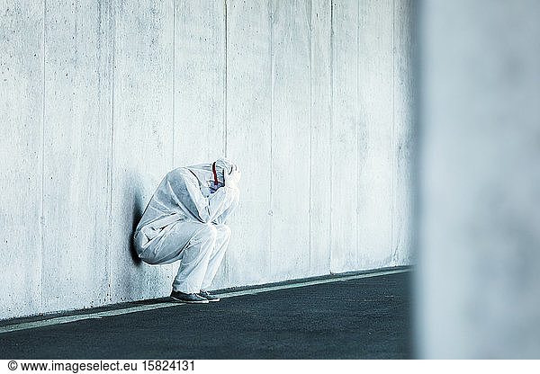 Verzweifelter Mann in Schutzkleidung lehnt an Betonwand