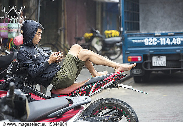 Vietnamese man relaxes on motorcycle and looks at smart phone.