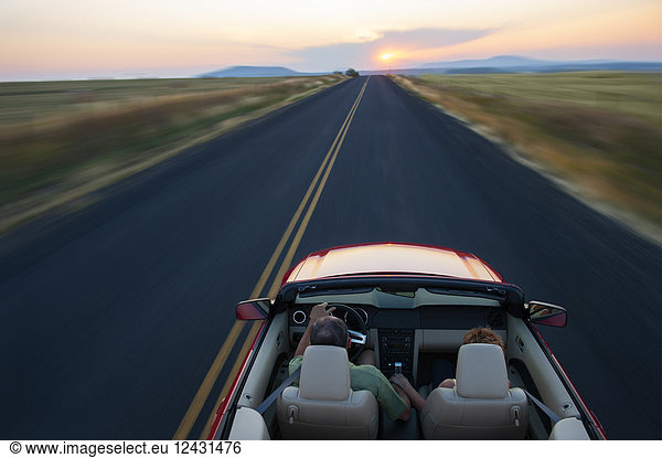 View from behind of senior couple in a convertible sports car driving on a highway at sunset in eastern Washington State  USA.