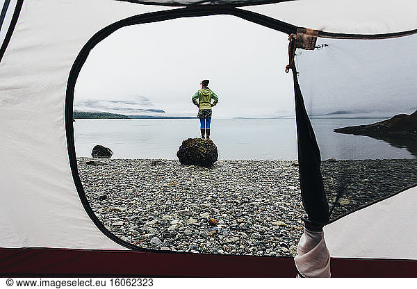 View through camping tent doorway of woman standing on beachan inlet on the Alaska coastline.