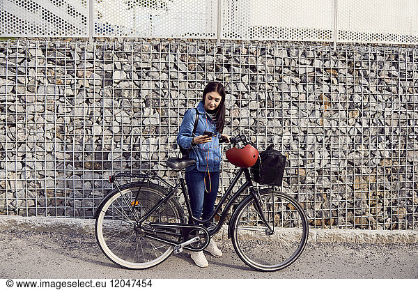 Volle Länge der Frau mit Smartphone beim Stehen mit dem Fahrrad an der Steinwand