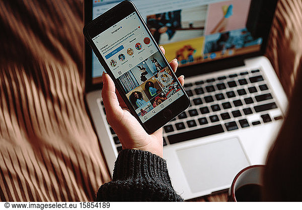 VSETIN  CZECH REPUBLIC/28 October 2019: Beautiful young girl sitting on the bed with laptop with cup of coffee and watching instagram on the screen of her smartphone. Winter  cozy  clothes an