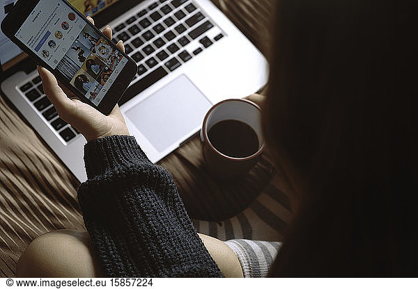 VSETIN  TSCHECHISCHE REPUBLIK/28. Oktober 2019: Junges Mädchen in Wintersocken sitzt mit Laptop auf dem Bett  hält eine Tasse Kaffee in der Hand und schaut auf ihrem Smartphone das Programm an. Winter  kuschelig  Kleidung und li