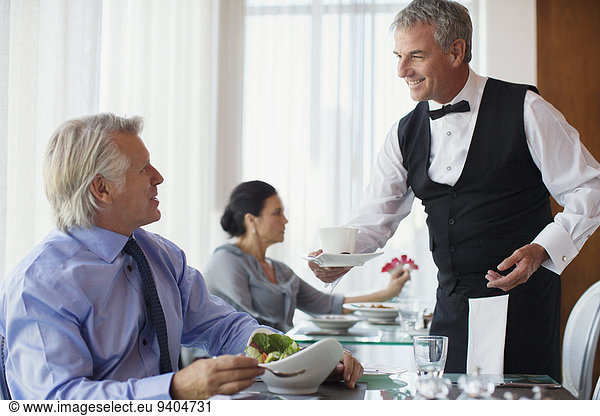 Waiter serving cup of coffee to mature man sitting at table in restaurant