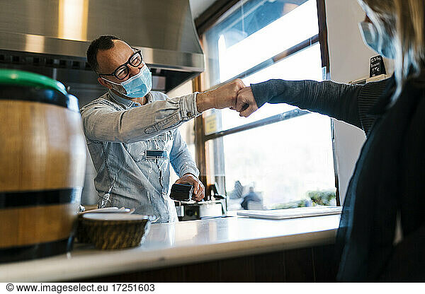 Waiter with protective face mask giving fist bump to female customer at a bar