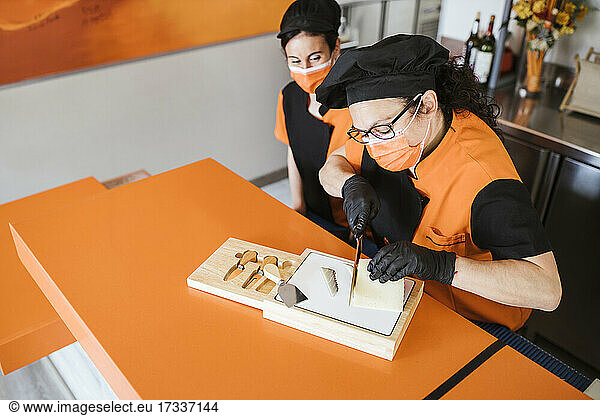Waitress looking at female chef cutting cheese on board during COVID-19
