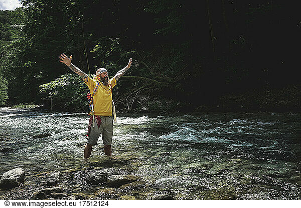 Walker with full beard and yellow hoodie standing in mountain river and raising arms