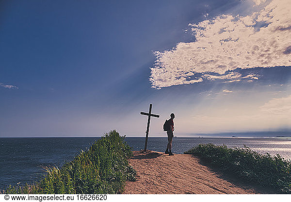 Wanderer am Gipfelkreuz stehend mit Blick auf das Meer gegen den Himmel bei Sonnenuntergang