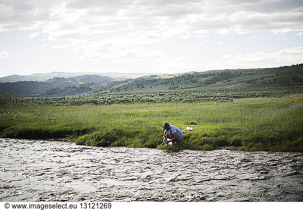 Wanderer füllt Wasserflasche  während er am Fluss vor bewölktem Himmel kauert