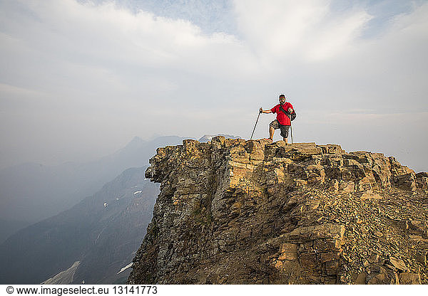 Wanderer in voller Länge mit Wanderstöcken auf Klippe gegen Berge stehend und bewölktem Himmel bei nebligem Wetter