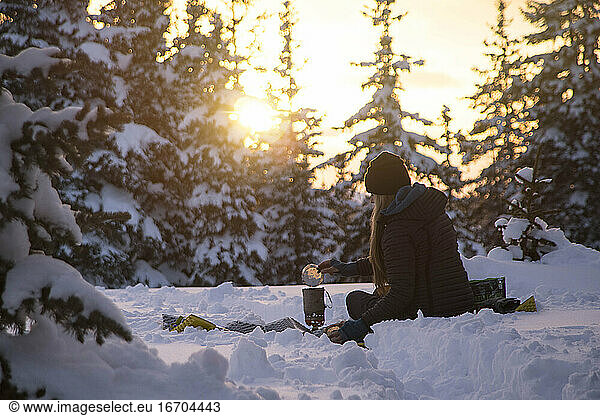 Wanderin bei der Essenszubereitung im Sitzen auf schneebedecktem Land bei Sonnenuntergang