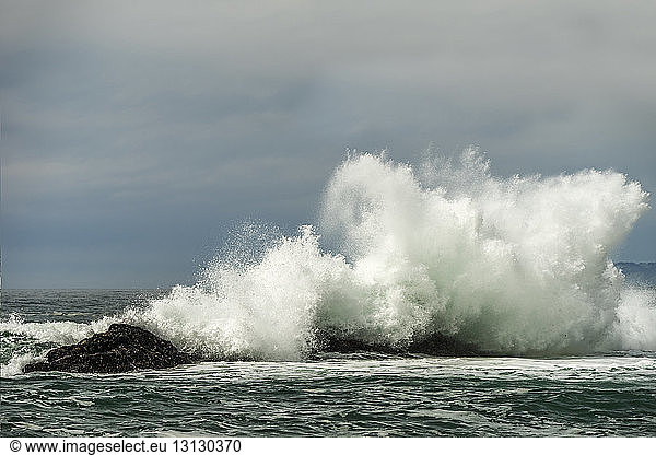 Waves splashing on rocks in sea against cloudy sky