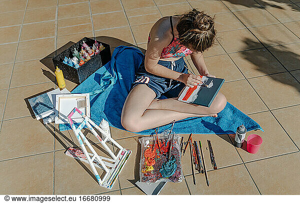 weißes Mädchen malt mit einem Pinsel ihr Bild auf einer Terrasse sitzend um Malerei Elemente. Overhead horizontales Foto