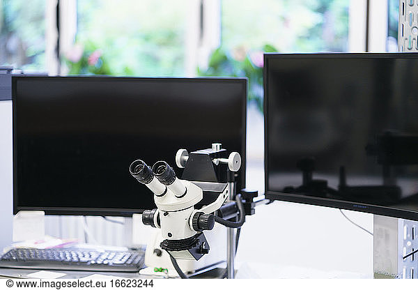 Weißes Mikroskop vor Computermonitoren in beleuchtetem Labor