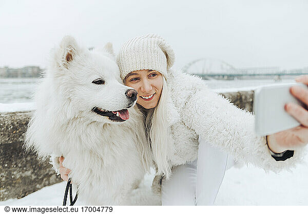 Weiß gekleidete Frau nimmt Selfie mit ihrem weißen Hund im Winter