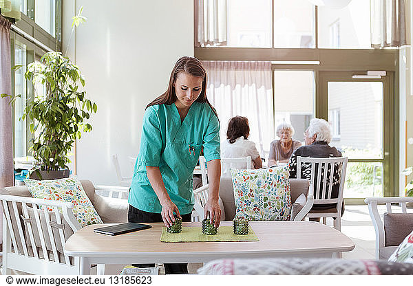 Weibliche Betreuerin arrangiert Tisch mit älteren Menschen im Hintergrund im Pflegeheim