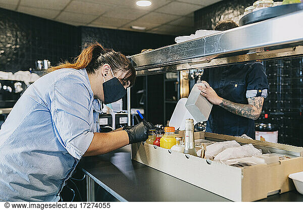 Weibliche Köchin  die auf eine Box zum Mitnehmen schreibt  während ihr Kollege an der Küchentheke im Restaurant steht